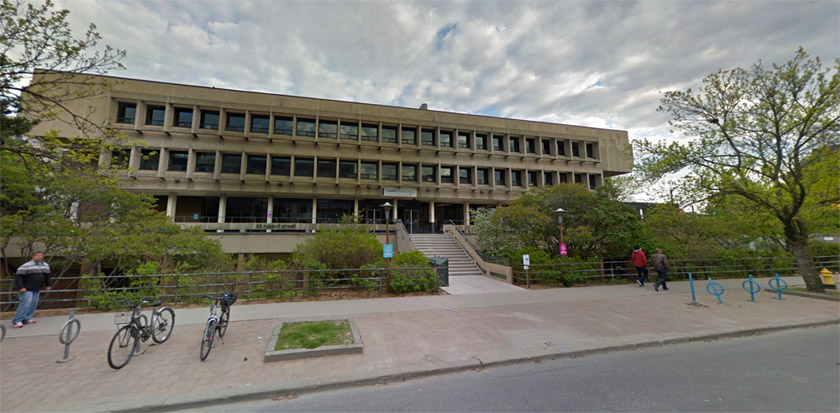 A wide, three-storey, brutalist building with a concrete staircase leading up to it from a wide sidewalk lined with bushes and trees.