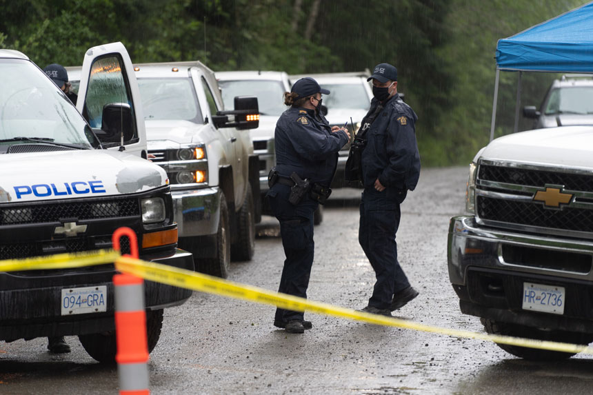 Two RCMP officers, a man and a woman, on a dirt road lined with police vehicles on each side. In the foreground is yellow police tape.