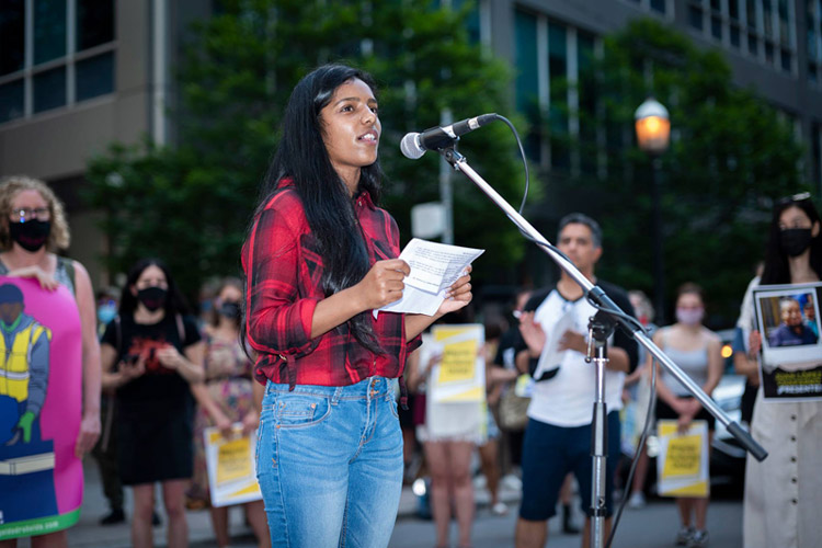 A young woman in a plaid red shirt and jeans speaks into a microphone set up on a street. Behind her are a row of people holding placards.