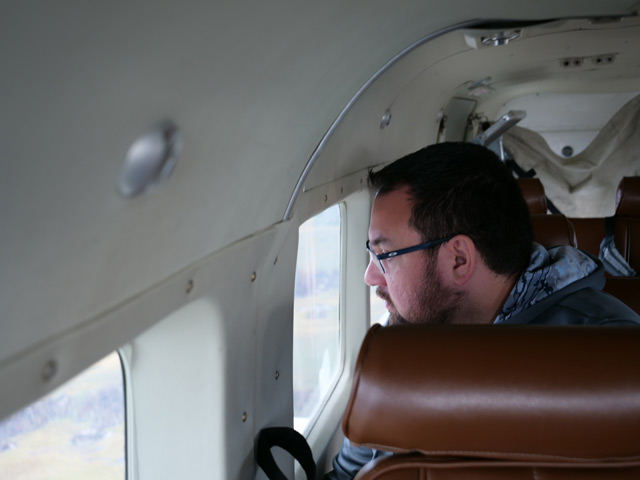 A man with glasses and a short-cropped beard gazes intently out of a passenger window of a small, likely propellor-driven airplane