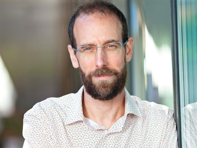 A man with a beard and glasses and tightly-patterned polka dot shirt, leaning up against a window and looking straight into the camera.