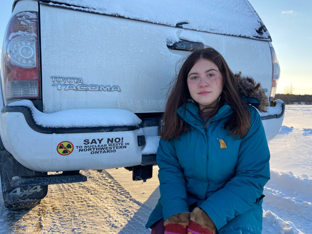 A young woman kneeling in the snow, next to the rear of a white Toyota Tacoma. There's a bumper sticker: SAY NO! to Nuclear Waste in Northwestern Ontario, with a radioactivity symbol with a red line through it.
