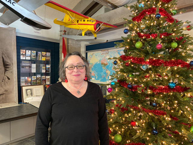 A somewhat older woman standing next to a brightly lit Christmas tree in what looks like some sort of museum but is probably actually the Ignace Town Hall. There are scale models of seaplanes hanging overhead.