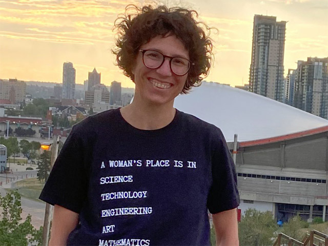 A woman photographed against a Calgary sunset, with the Saddledome not too far behind her. Her tshirt says A WOMAN'S PLACE IS IN: SCIENCE, TECHNOLOGY, ENGINEERING, ART, MATHEMATICS