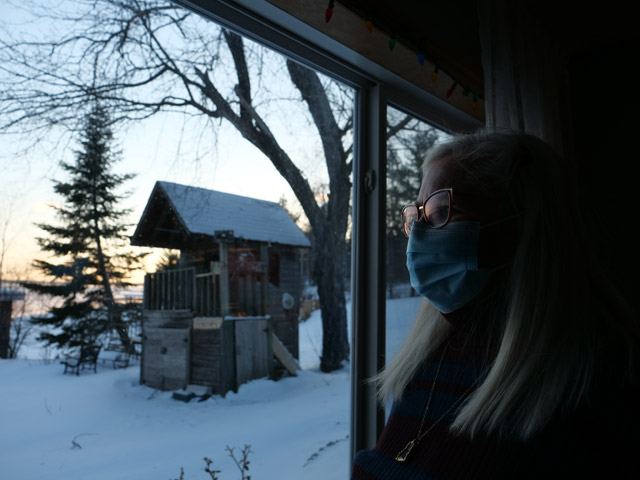 A photo of a woman in a surgical mask gazing out a window toward a snowy backyard at sunset. There appears to be a wooden treehouse or fort of some sort.
