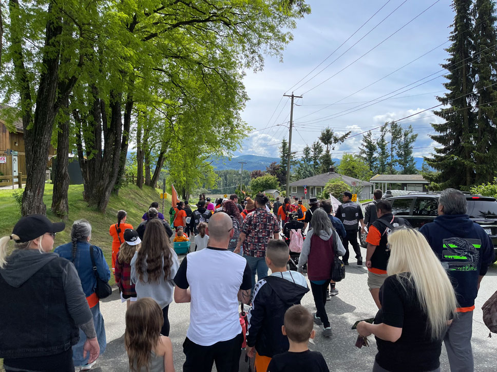 Several dozen people of varying ages, many in orange shirts, walking down a residential road lined with bright green trees. There's a grand, mountainous vista in the distance in front of them.