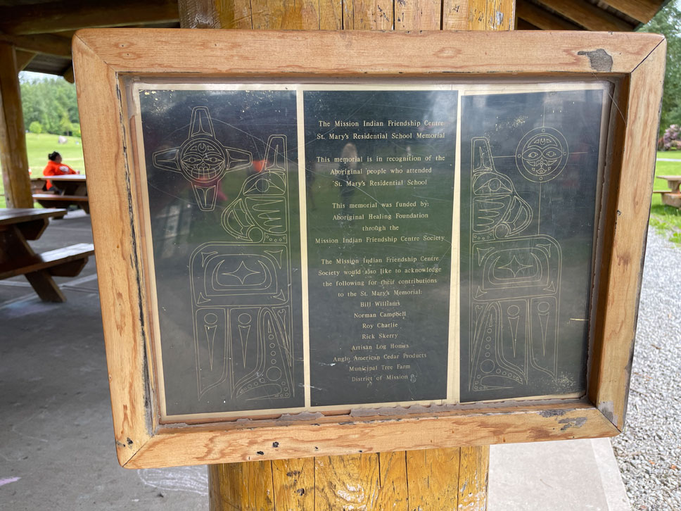 A black plaque, with images and words etched into it, inside a wooden frame, affixed to a supporting column of a large wooden structure covering a picnic area.